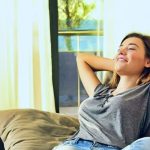 Young woman on couch in calm room, enjoying natural light. Depicts comfort linked to HVAC services by SOS Heating & Cooling.