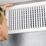 Individual inspecting a grilled air vent cover for HVAC maintenance in a home environment.