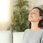 Woman sitting calmly in an indoor space with soft furnishings and a green plant, promoting relaxation and mindfulness.